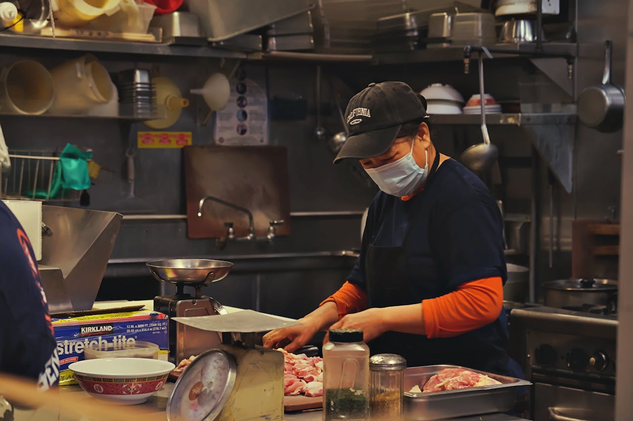 A woman in a kitchen wears a face mask while cutting meat, showcasing food preparation safety.